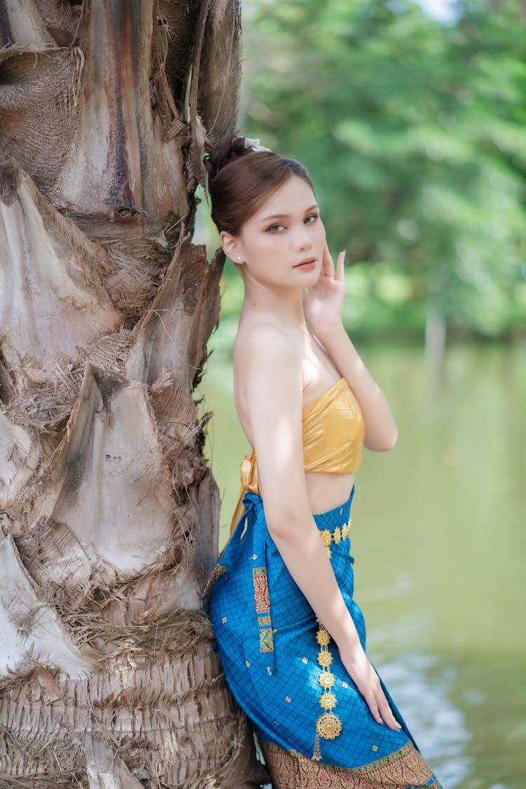 Model In A Golden Tie Back Tube Top And A Printed Blue Skirt Standing Under A Palm Tree On The Riverbank