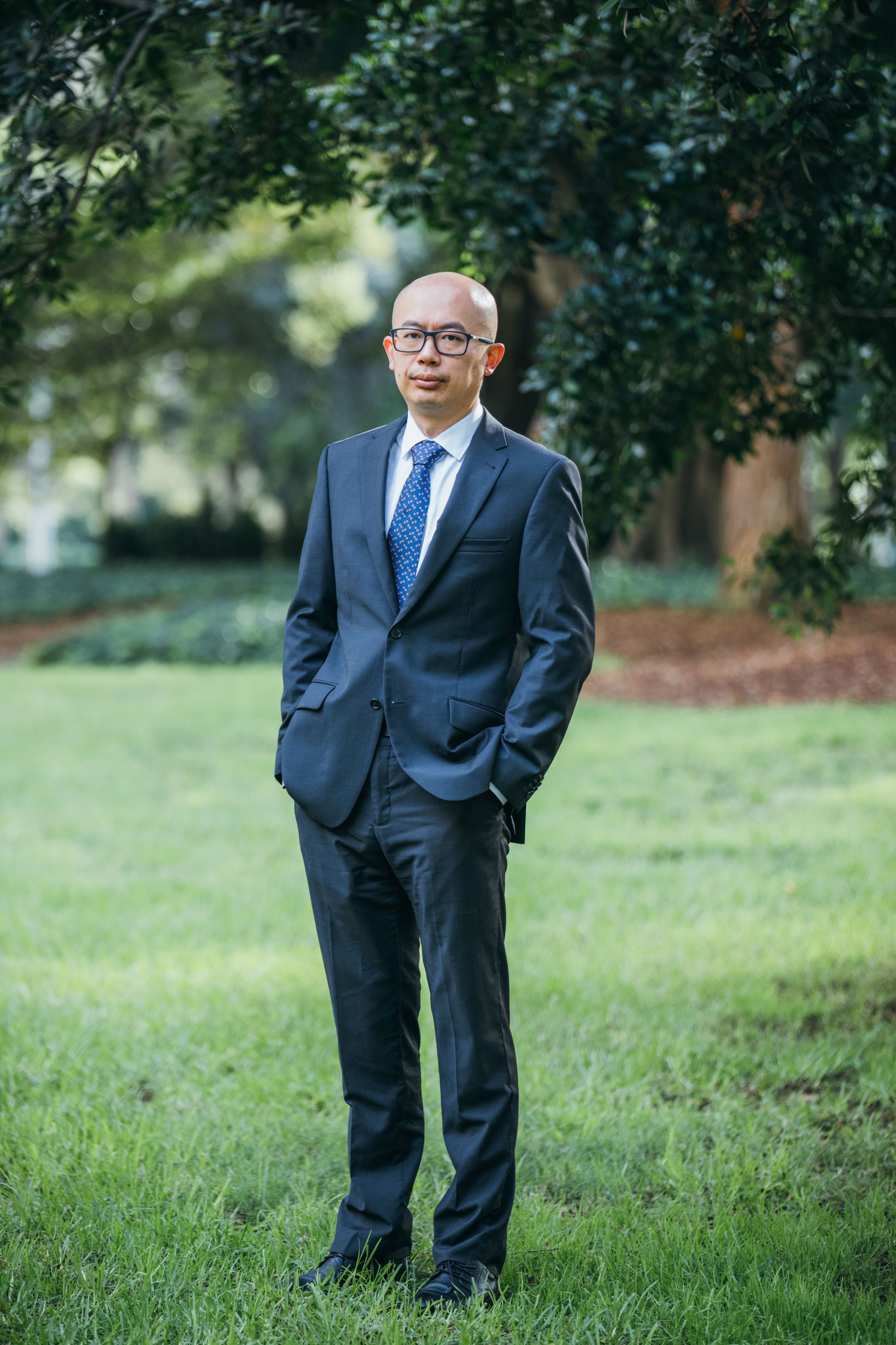 Bald Asian businessman in suit with glasses standing in a lush green park in Sydney, Australia.