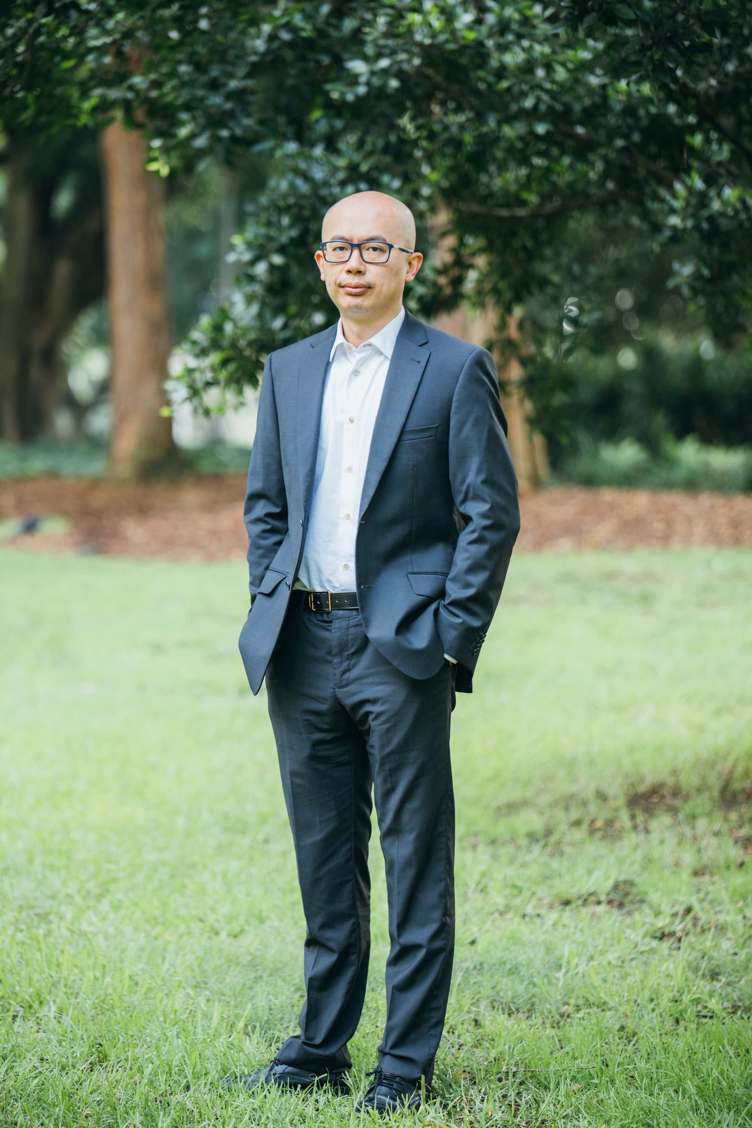 Bald Asian man in suit standing confidently outdoors in Sydney park.