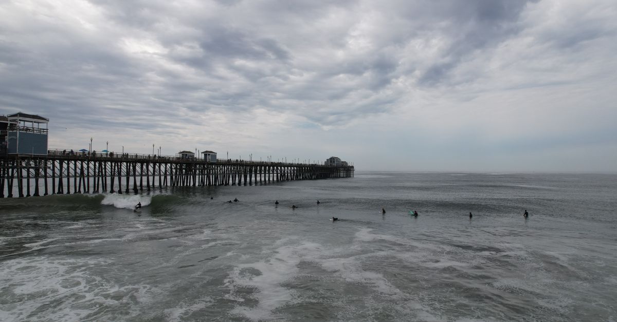 Surfers ride waves near Oceanside Pier, California under a moody sky.