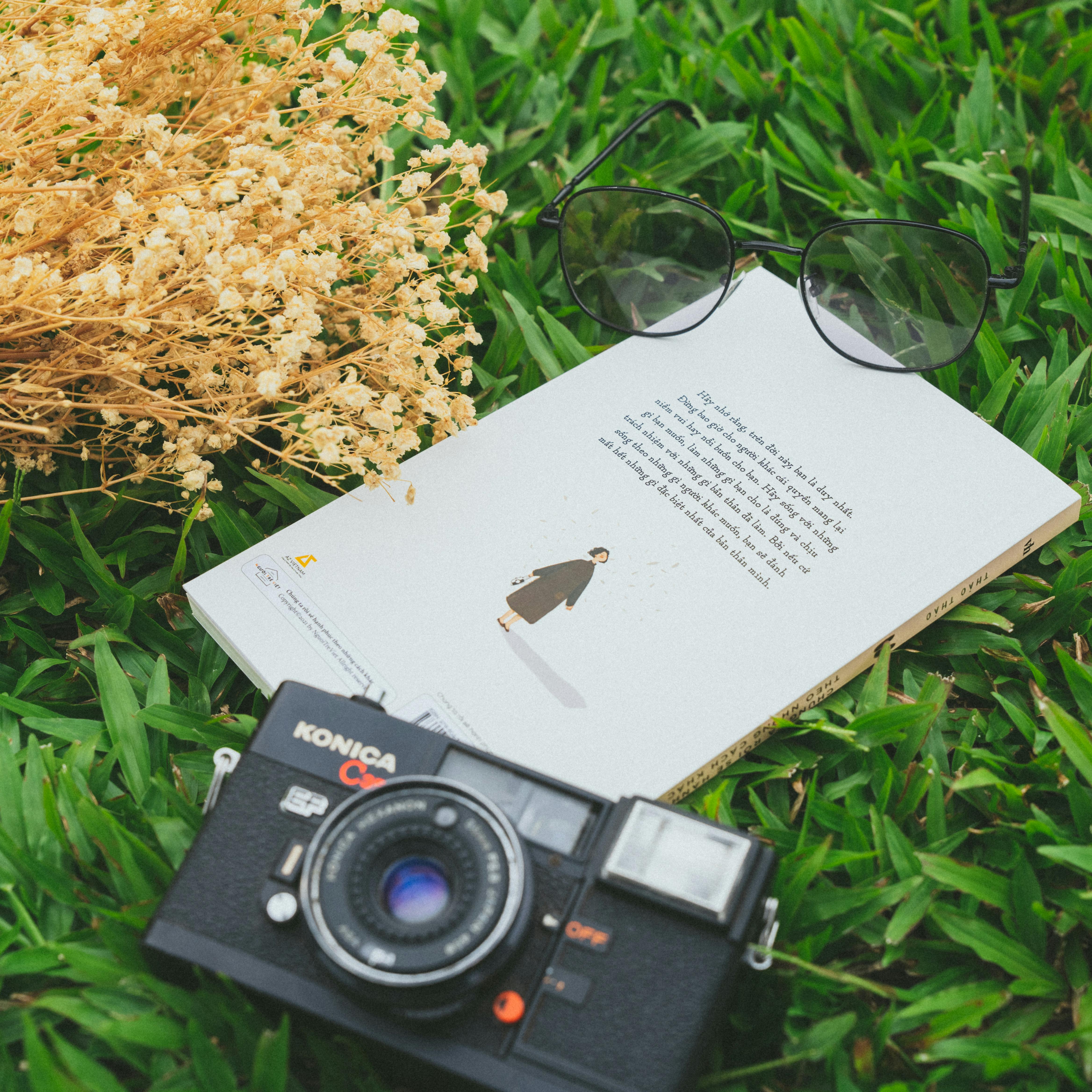 A vintage Konica camera with a book and sunglasses on a grassy outdoor setup.