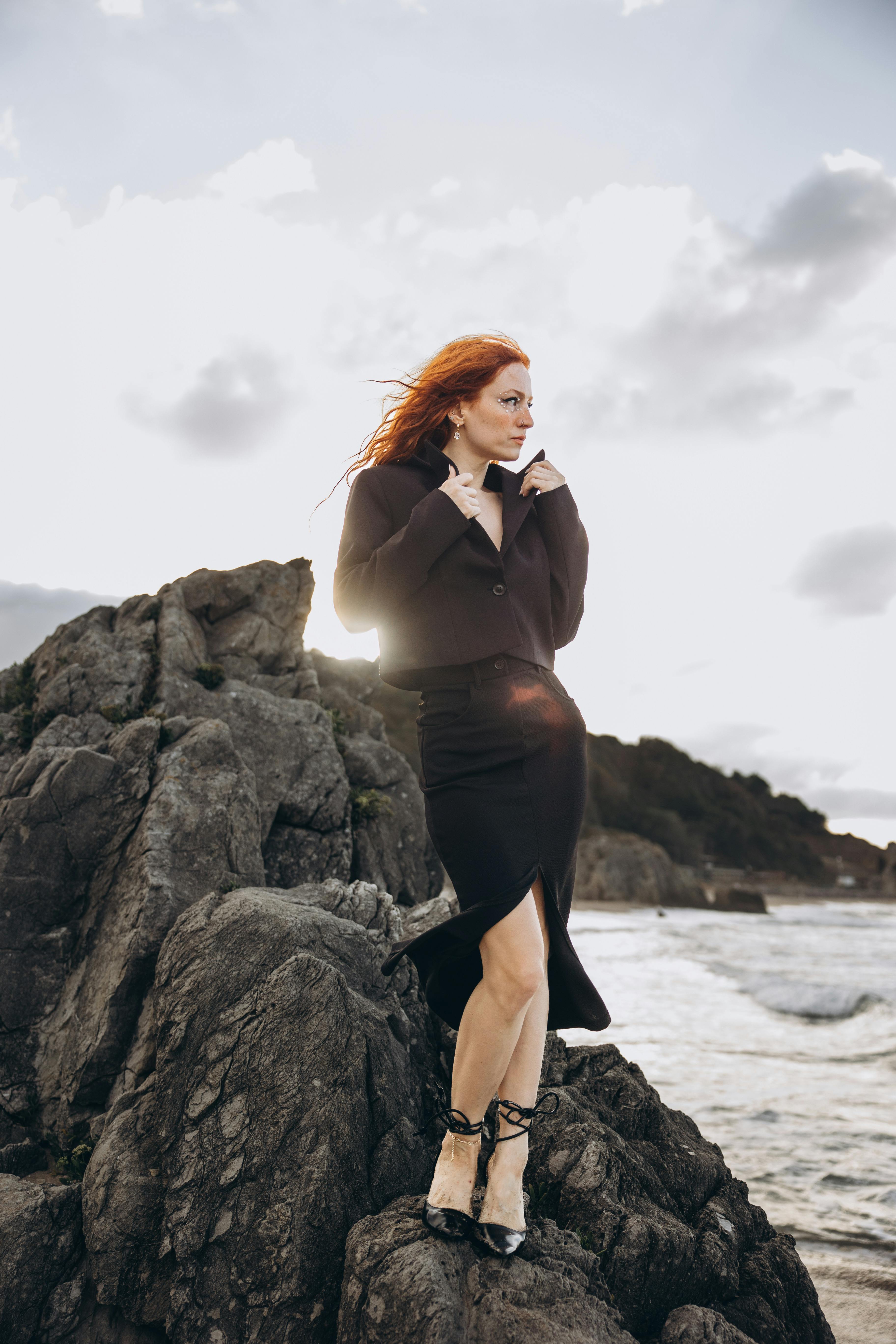 a woman with red hair standing on rocks near the ocean