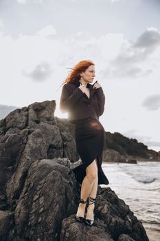 Stylish woman with red hair stands elegantly on a rocky beach in Türkiye, exuding confidence.