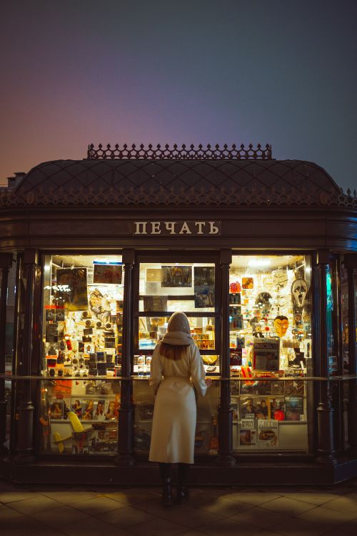 Free A woman stands in front of an illuminated newsstand at night. Stock Photo