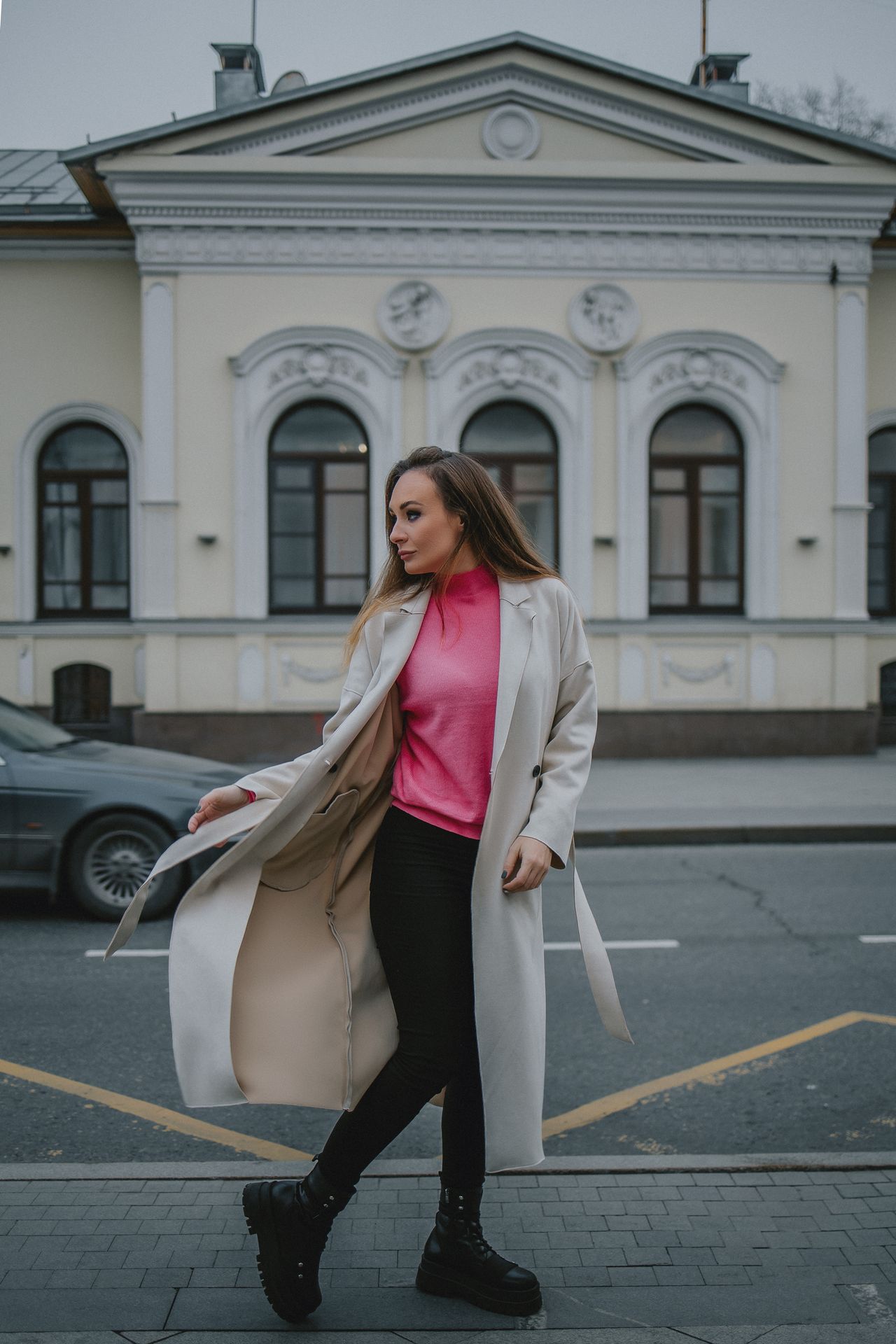 Woman in a cream coat, pink sweater, and black boots on a city street