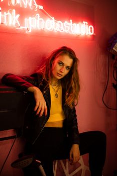 Fashionable woman in a leather jacket poses under neon light in an edgy studio setting.