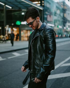A fashionable man wearing sunglasses strolls through urban streets in a black leather jacket, capturing modern city style.