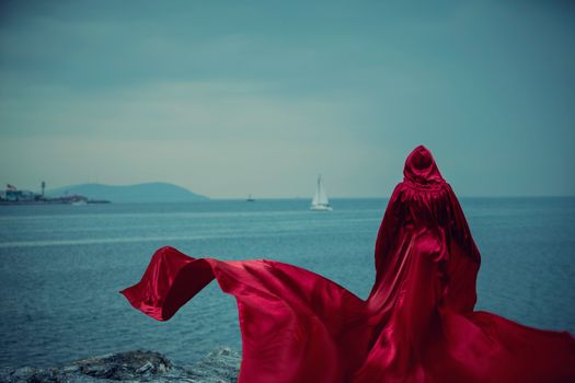A woman with a red cloak stands on a cliff, gazing at the distant sailboat over the ocean.