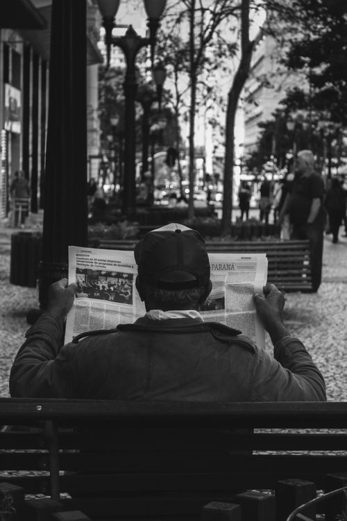 Free A man sits on a bench in an urban park, reading a newspaper, captured in a black and white photo. Stock Photo