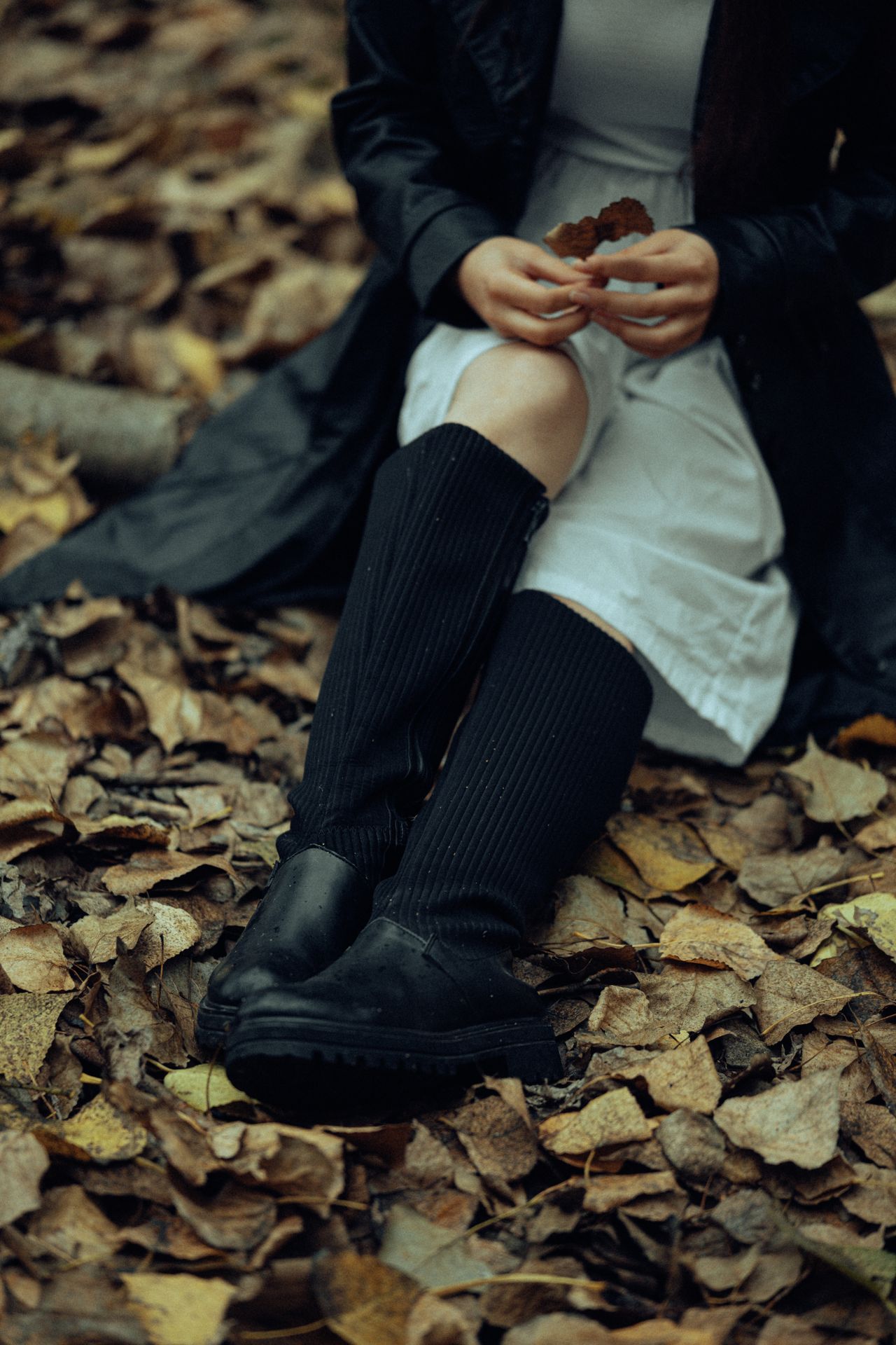 Woman in a white knit dress and black ribbed knee-high boots sitting among autumn leaves