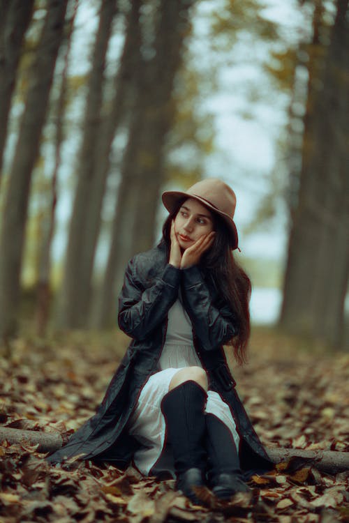 Woman in Black Coat and Hat Sitting with Hands on Cheeks on Autumn Leaves