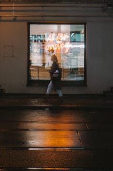 A woman walks past a neon-lit shop window on a rainy Berlin street, reflecting urban vibes.