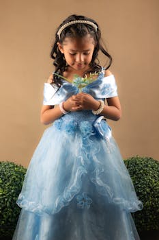A young girl in a lacy blue dress smiling and holding a flower in a studio shoot.