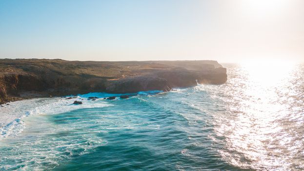 Aerial shot of sunlit cliffs and ocean waves reflecting light, showcasing coastal beauty.