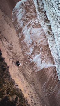 Dynamic aerial shot of waves crashing on a sandy shore with visible tire tracks.