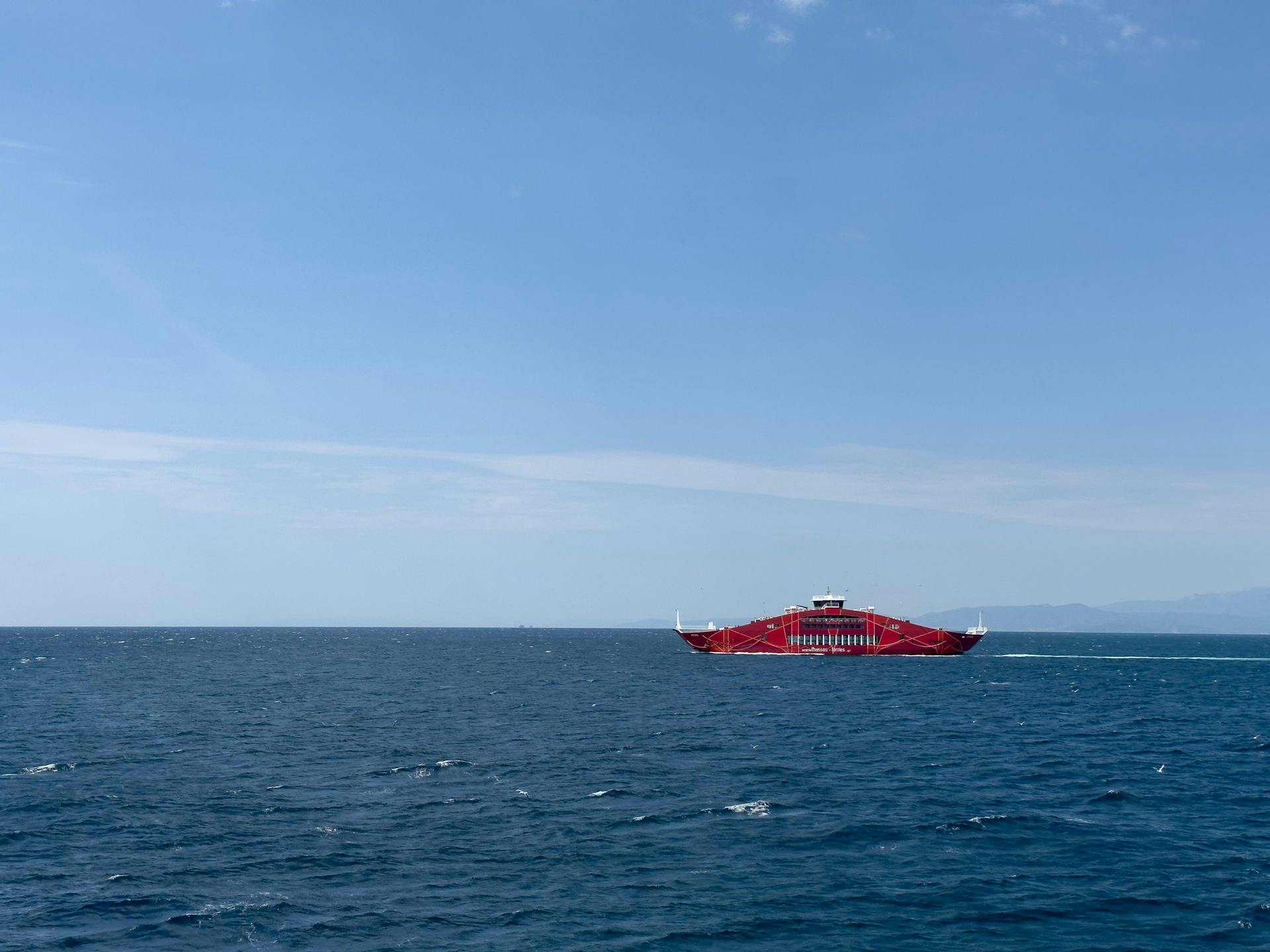 A vibrant red ferry navigates the blue Aegean Sea near Keramoti, Greece under a clear sky.