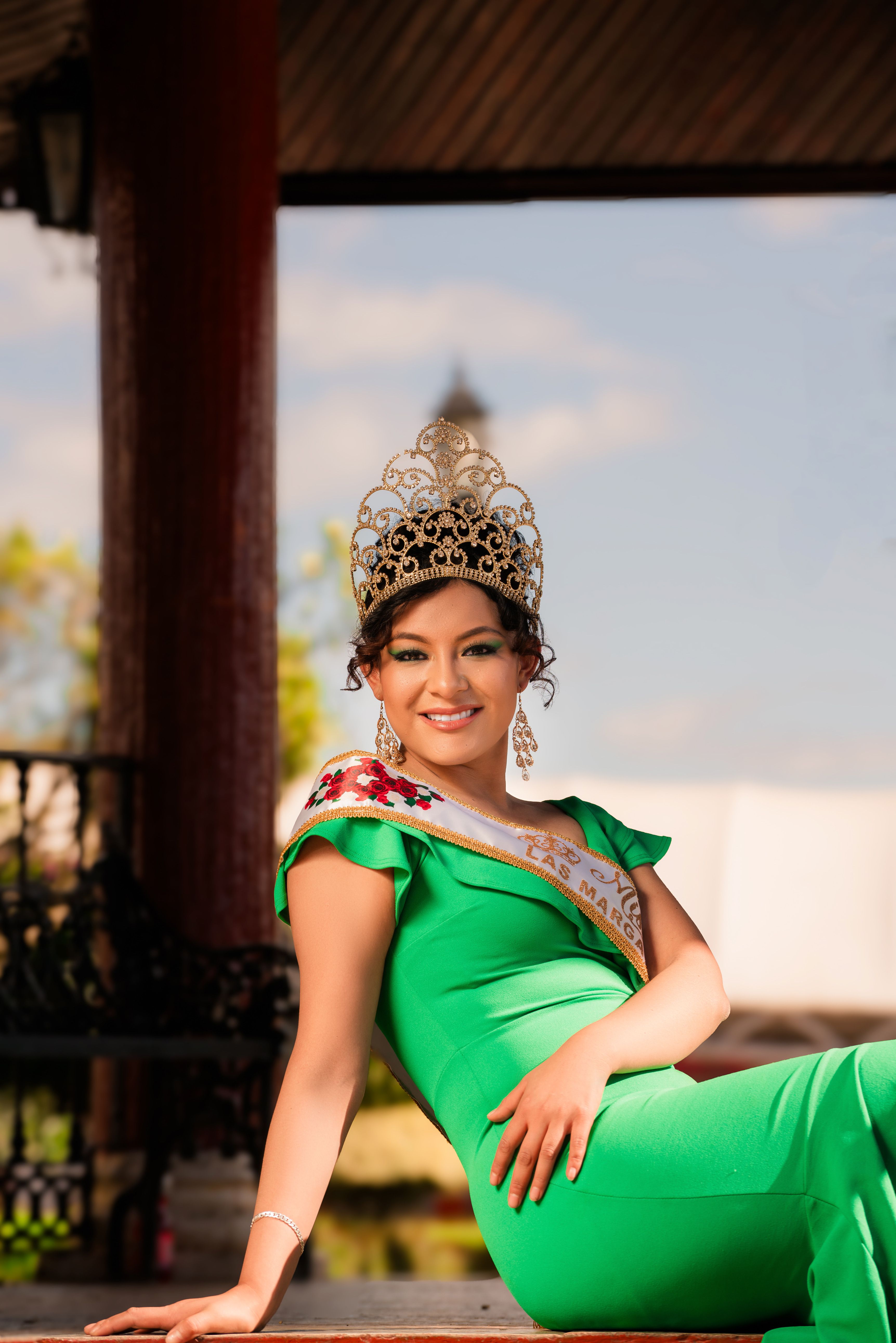 A woman wearing a green dress, tiara, and sash, smiling elegantly in an outdoor setting.