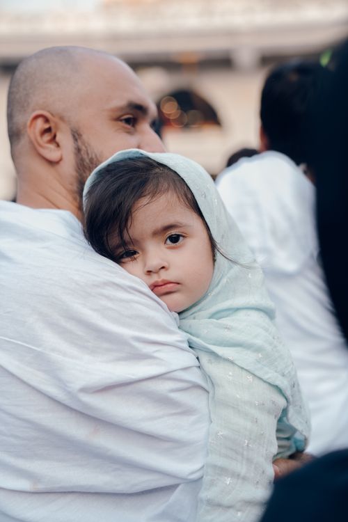 Free A father carrying his young daughter wrapped in fabric during an outdoor event, both wearing white attire. Stock Photo