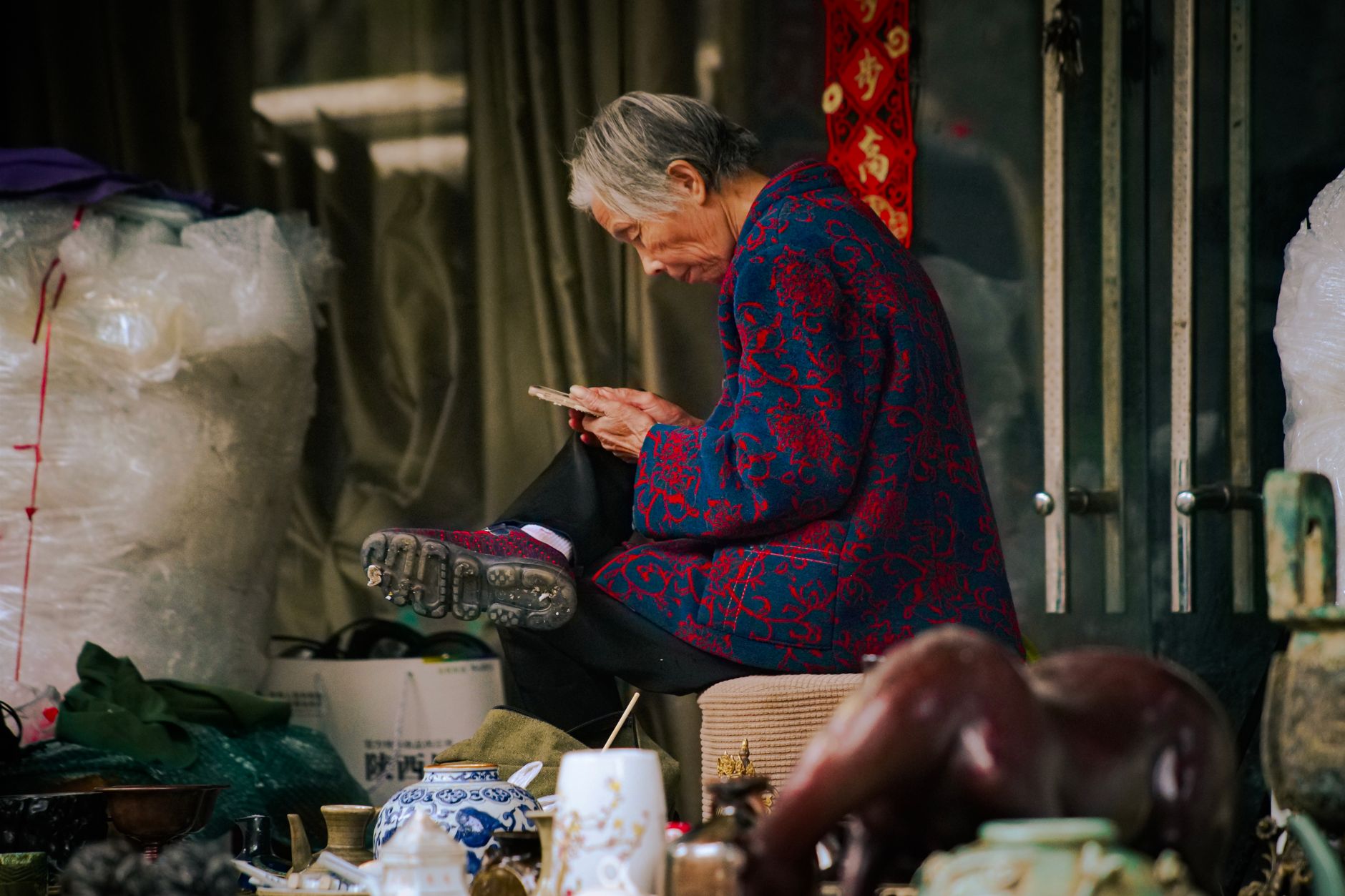 Elderly woman in traditional jacket, seated at outdoor market, browsing smartphone.