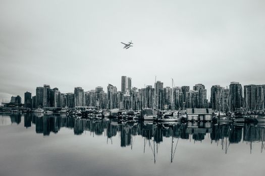 Monochrome Vancouver cityscape with seaplane over marina, showcasing reflective skyline.