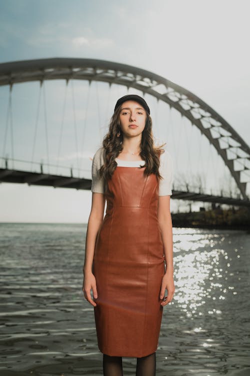 A woman in a brown leather dress stands by the water