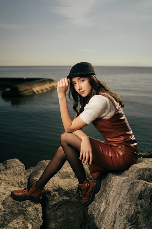 A woman in a leather dress and hat sitting on rocks by the water