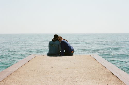 Free A young couple sitting affectionately on a pier by the sea, enjoying a sunny day. Stock Photo