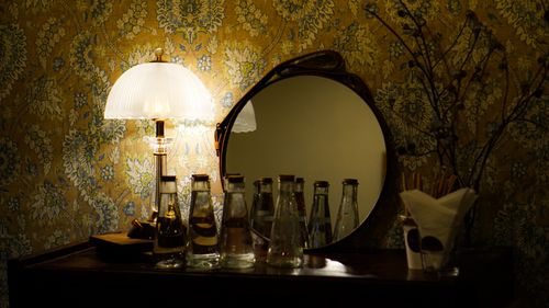 Free A vintage-style setup featuring a decorative lamp, bottles, and a mirror reflecting elegant wallpaper. Stock Photo