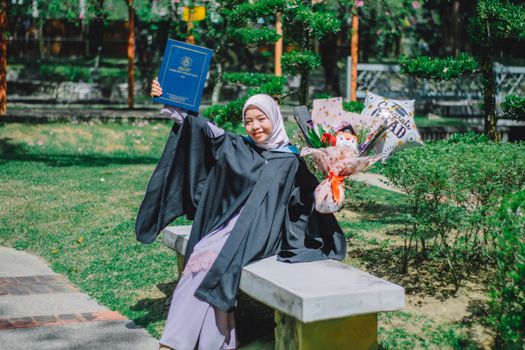 Happy woman in graduation gown sitting on a bench, celebrating in Shah Alam, Malaysia.