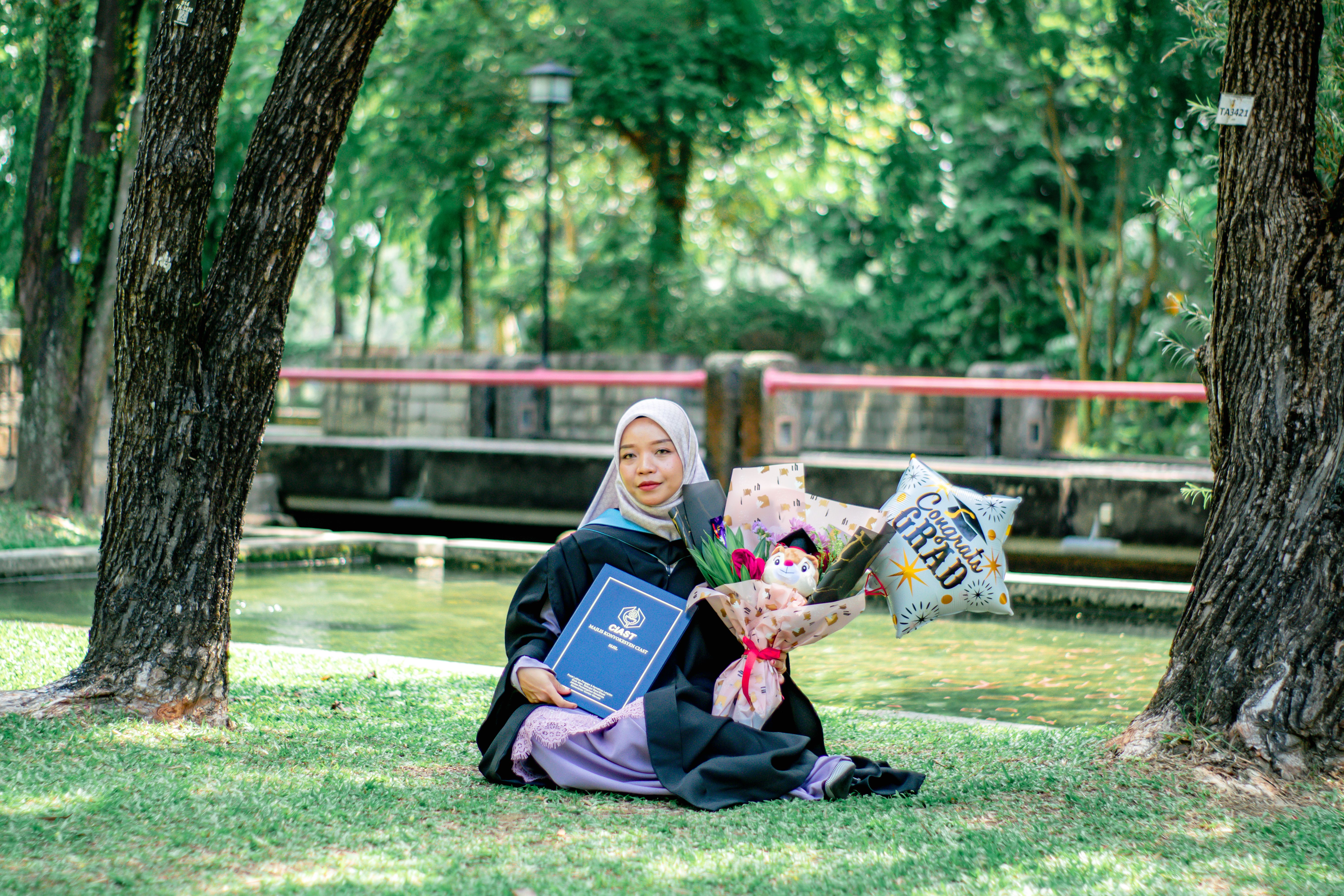 Young woman celebrates graduation in Shah Alam park with diploma and flowers.