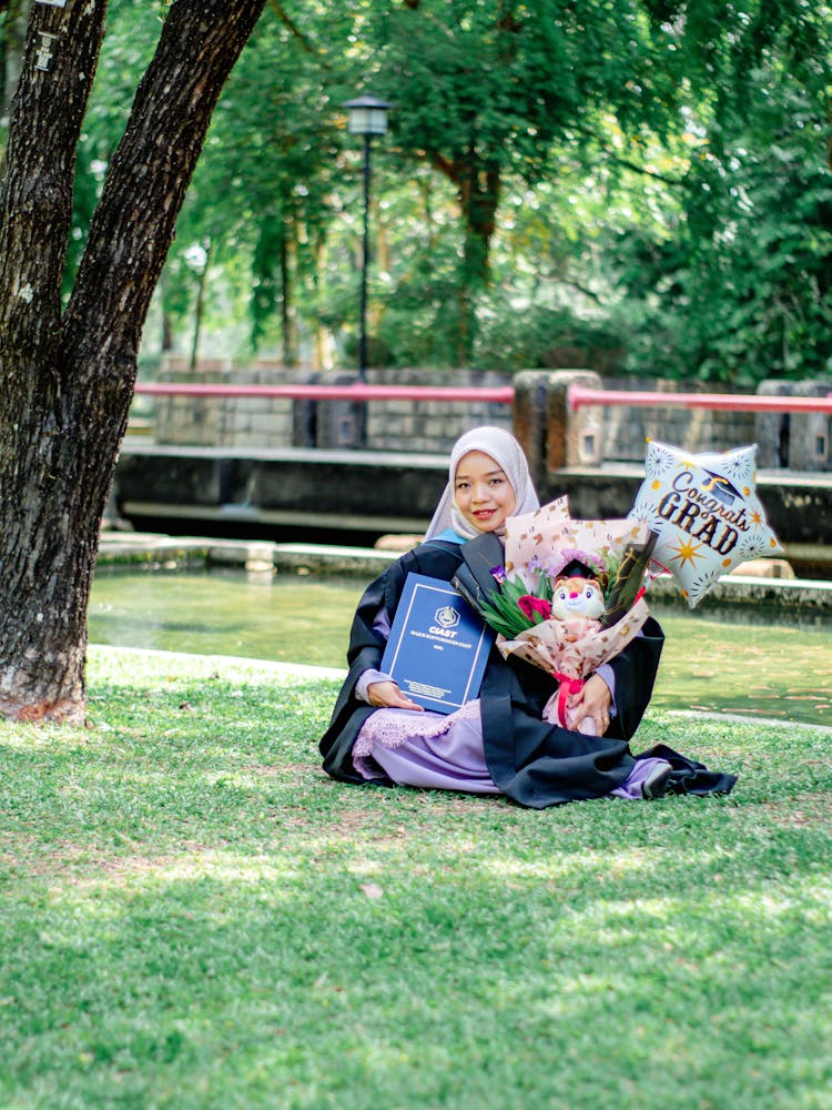 A Girl In A Graduation Gown Sitting On The Grass In A Park With Flowers And Balloons 