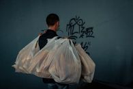 A man carries plastic bags full of groceries past a graffiti-covered wall.