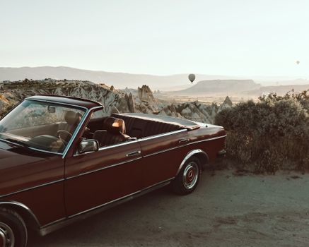 A vintage convertible car parked on a hill with hot air balloons in the background during sunset.