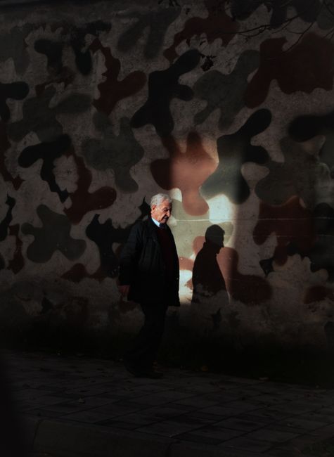 An elderly man walks by a camouflage-painted wall in Tirana, Albania, casting a shadow in evening light.