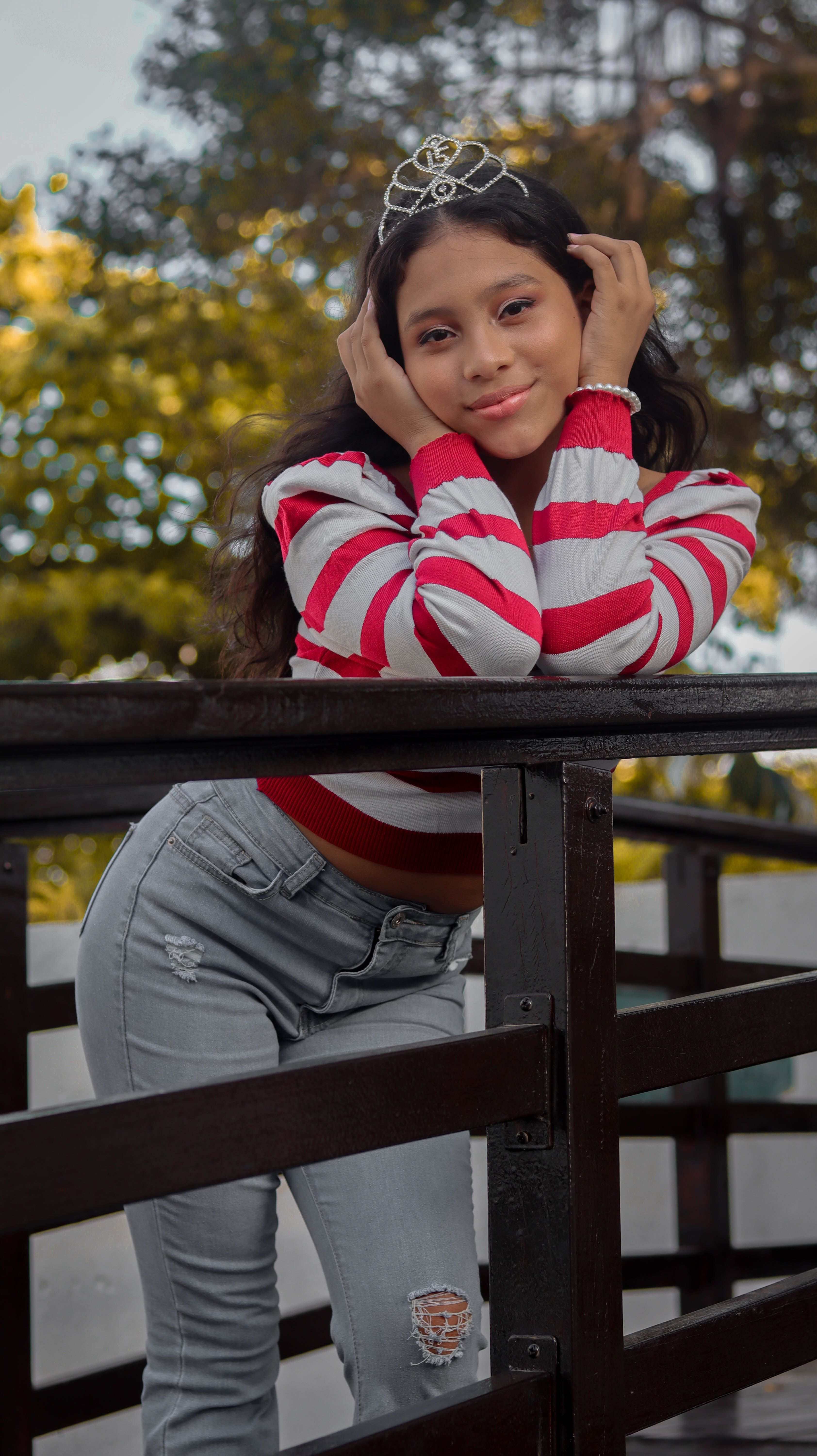 Young woman in striped shirt and jeans, wearing a tiara, standing outdoors and smiling.