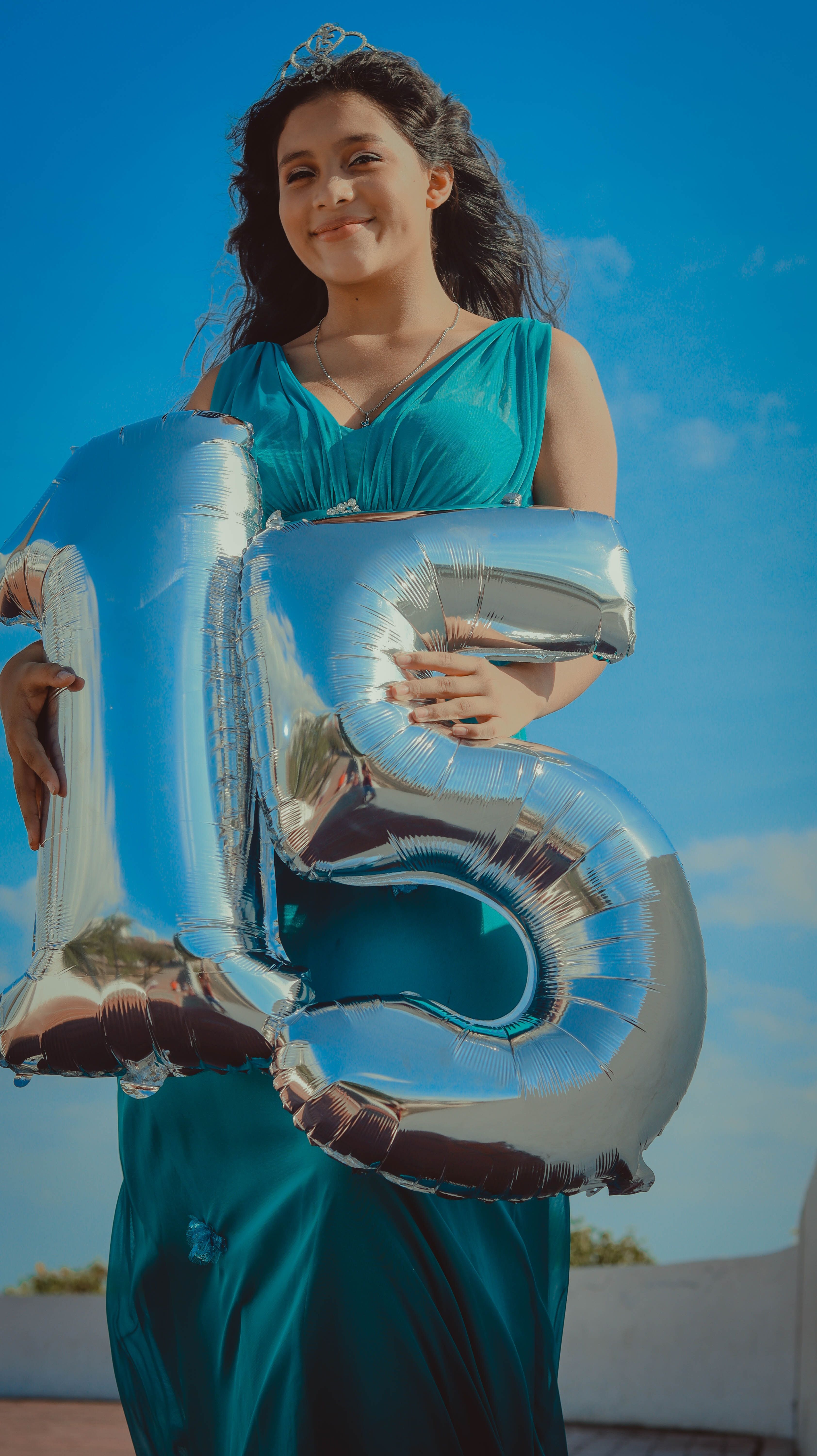 Smiling teen girl holding '15' balloons in celebration, outdoors under a blue sky