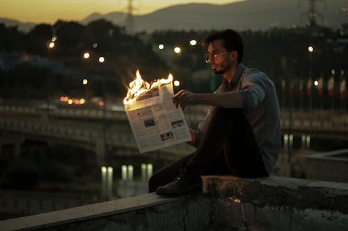 Man Sitting on a Wall Outside and Burning a Newspaper 