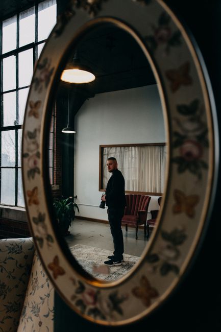A photographer stands inside a modern room reflected in a decorative mirror.