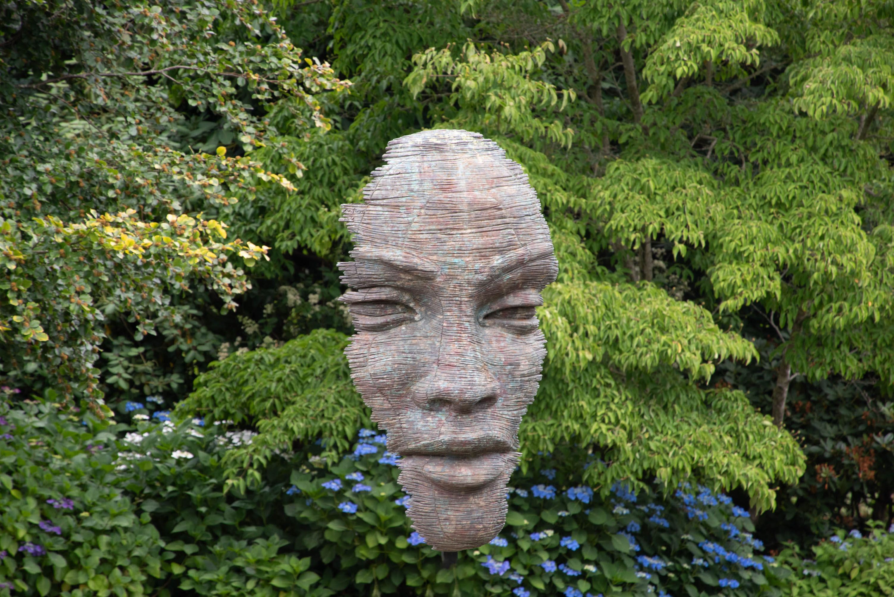 Artistic face sculpture in vibrant garden surrounded by green foliage and flowers.
