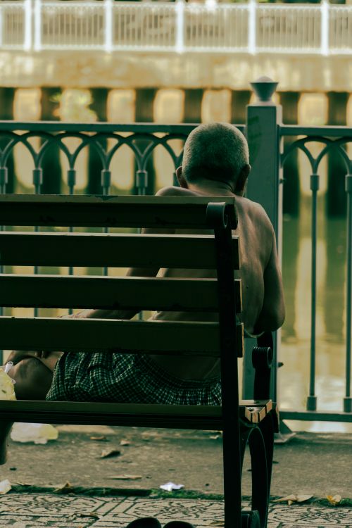 Free A shirtless man sits alone on a bench by a canal, facing away. Stock Photo