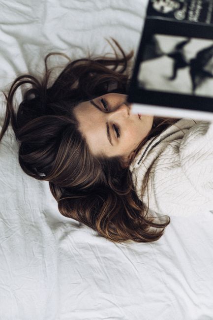 A young woman with long hair lying on a bed while reading a book, capturing a relaxing moment.