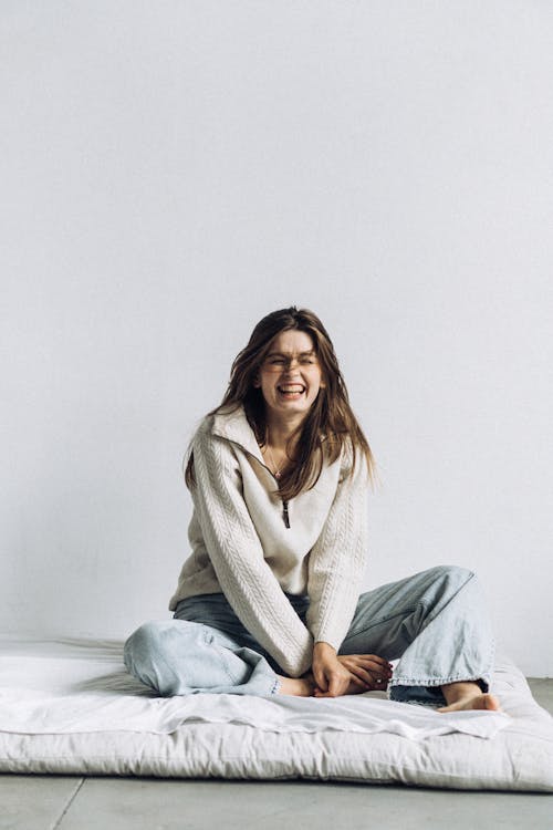 Laughing Young Woman in Sweater and Jeans Sitting on a Mattress on the Floor