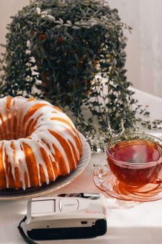 A cozy vintage setup featuring a bundt cake with icing, a cup of tea, and a classic camera.