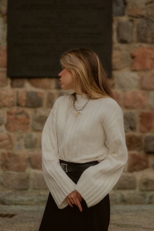 Free Fashionable woman in a white sweater stands outdoors against a rustic stone wall. Stock Photo