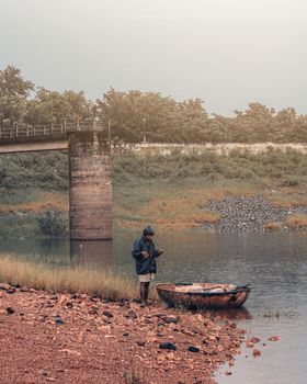 A serene scene of a man by a riverbank with a small boat, bridge, and mist in the morning air.