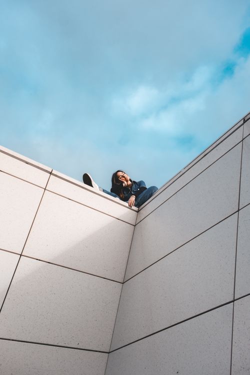 Free A woman in casual attire sits on the edge of a modern building against a blue sky. Stock Photo