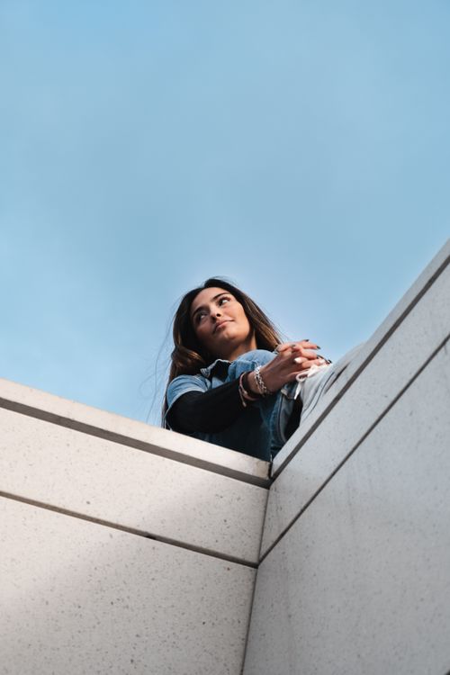 Free Young woman sitting on a rooftop ledge under a clear blue sky, reflecting and relaxing. Stock Photo