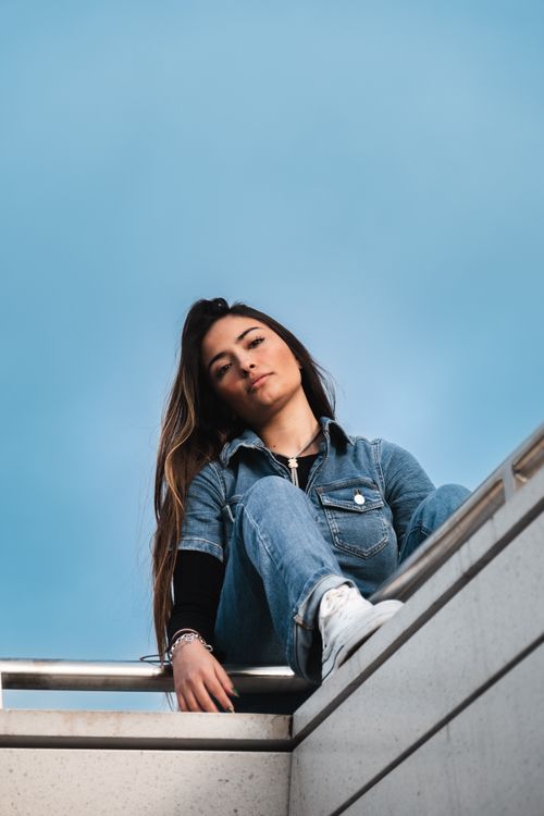Free Stylish urban portrait of a woman in a denim outfit, sitting outdoors against a clear sky. Stock Photo