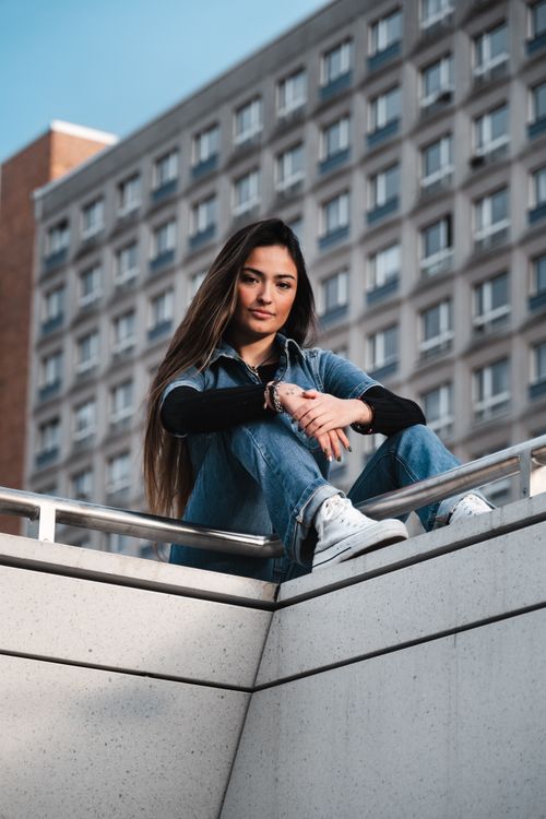 Free A stylish woman smiling while sitting on a building ledge in a city setting, wearing casual denim. Stock Photo