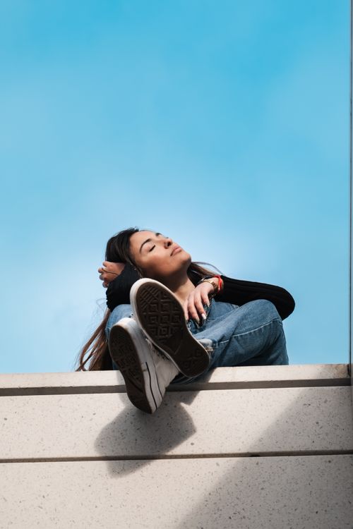 Free Young woman sitting on rooftop edge in denim against clear blue sky, enjoying sunlight. Stock Photo